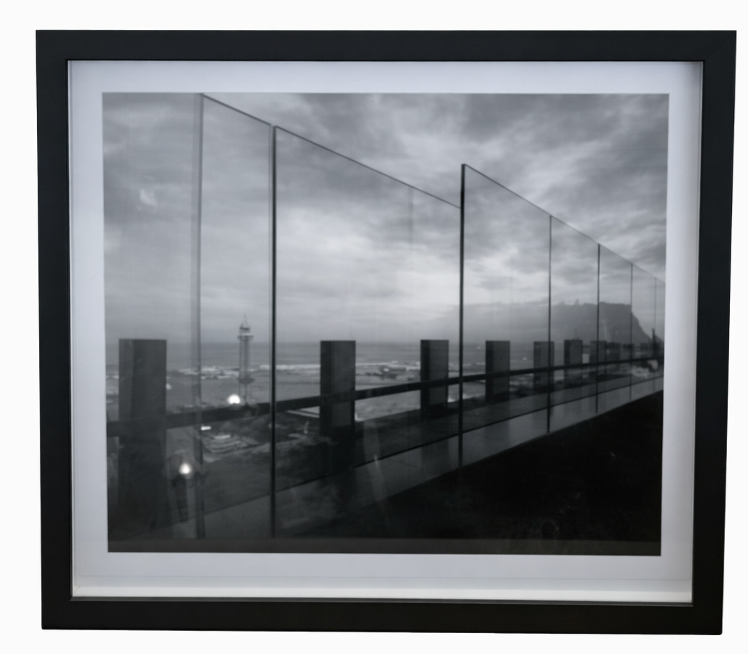 Black and white framed painting by Delphine Dumas depicting a modern glass balcony overlooking a coastal scene with a lighthouse and cloudy sky.