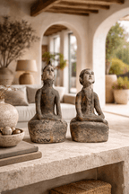A pair of aged stone-like seated sculptures on a light stone table with a blurred outdoor patio in the background.