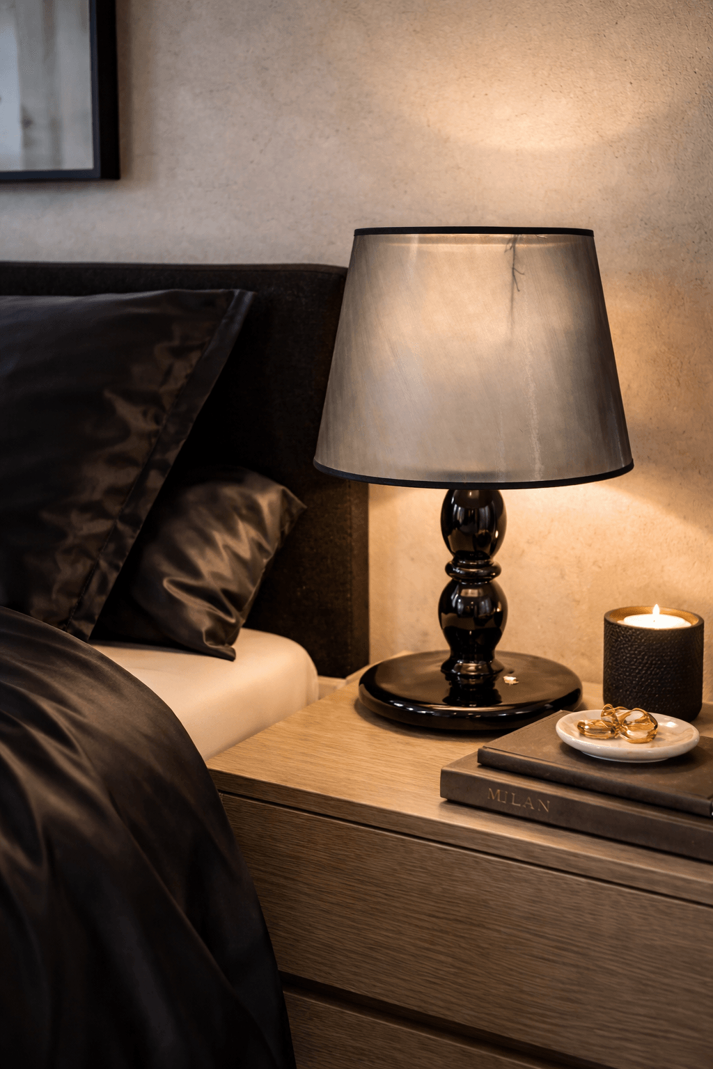 Black table lamp with a dark gray translucent shade, on a wooden nightstand beside a bed with satin bedding.