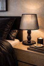 Black table lamp with a dark gray translucent shade, on a wooden nightstand beside a bed with satin bedding.