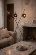 Neruda Floor Lamp with a dark metal cross-legged frame and three smoky glass globes emitting a warm glow in a living room.