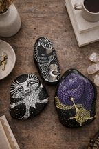 Black stones hand-painted with creature and spiral designs, displayed on a rustic wooden table.