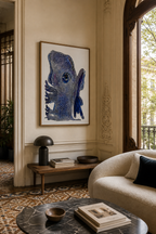 Blue Entity artwork in a wooden frame on a beige wall, above a dark mushroom lamp and books.