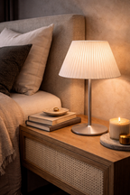 White pleated table lamp by Creditia with a sleek silver base on a wooden nightstand with books and a candle.