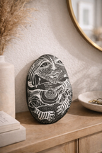 Black and white art stone featuring intricate line illustrations of a face and hands, leaning upright on a wooden cabinet.