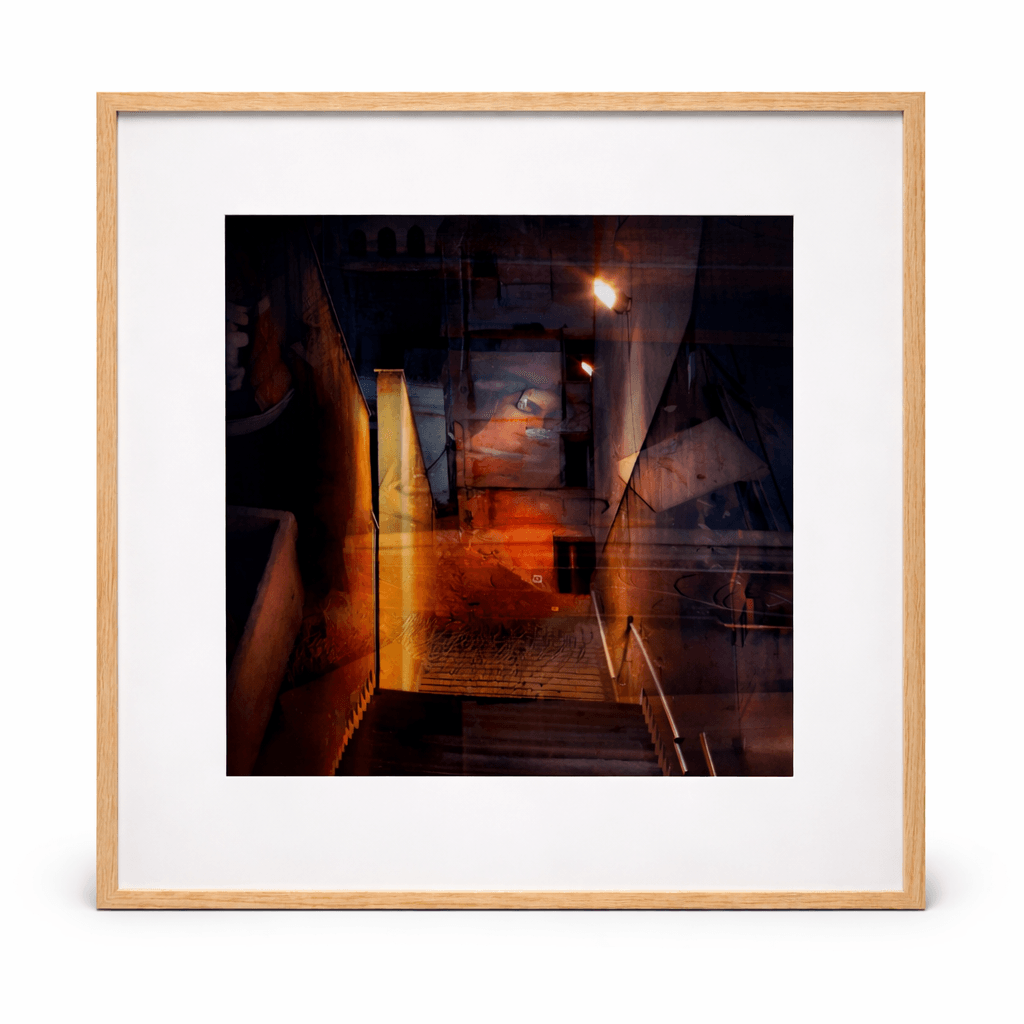 Framed painting titled Escala by Manuel Cohen, showing a layered dark urban scene with orange streetlights and a staircase, in a wood frame.