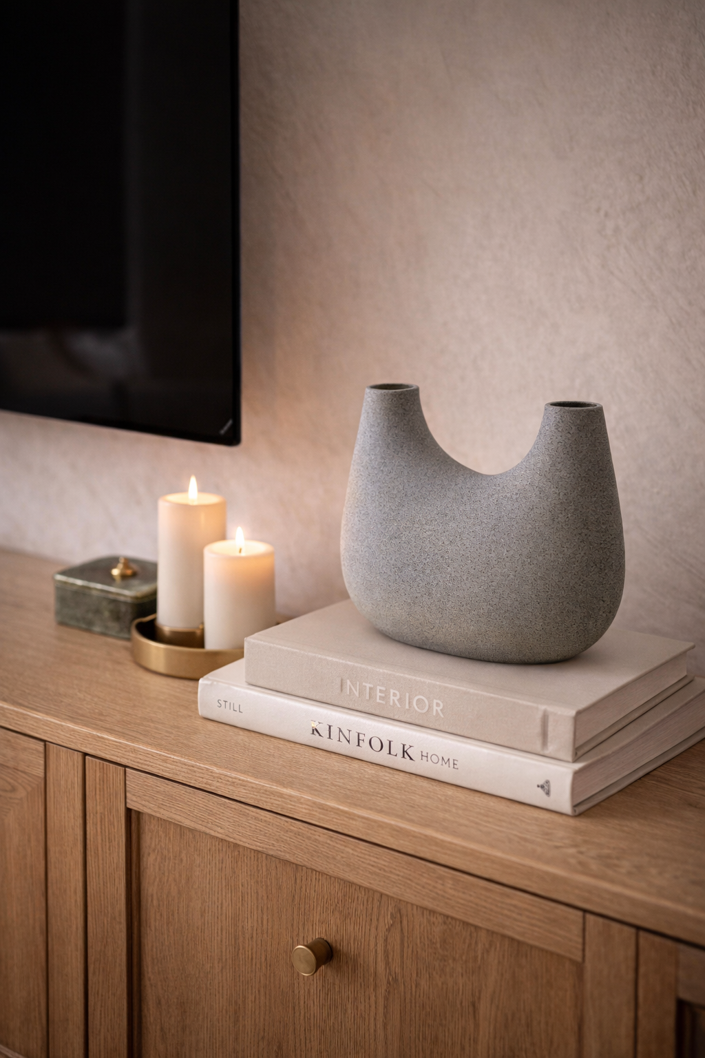 Small speckled grey U-shaped vase sitting on a stack of books on a wooden cabinet next to lit candles.