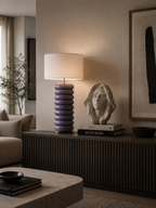 Purple stacked ring table lamp with a white drum shade, illuminated on a dark wooden console table.