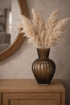 Brown ribbed glass vase holding dried pampas grass, displayed on a wooden cabinet.