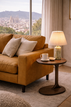 Dark brown wood Fenix Side Table with a round top and sculptural pedestal base, positioned beside a camel sofa.