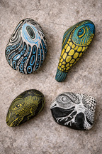 Four pieces of Eagle Stone Art, hand-painted with black, white, blue, and yellow abstract and animal-inspired designs, on a gray stone surface.