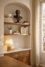 Bronze bust sculpture of a bearded man, displayed on a wooden shelf in an arched alcove with books, a plant, and a lamp.